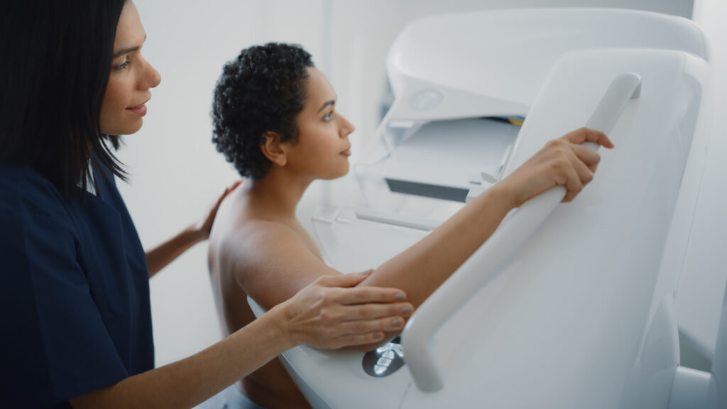 Woman holding mammogram gantry machine while mammography technologists helps her.